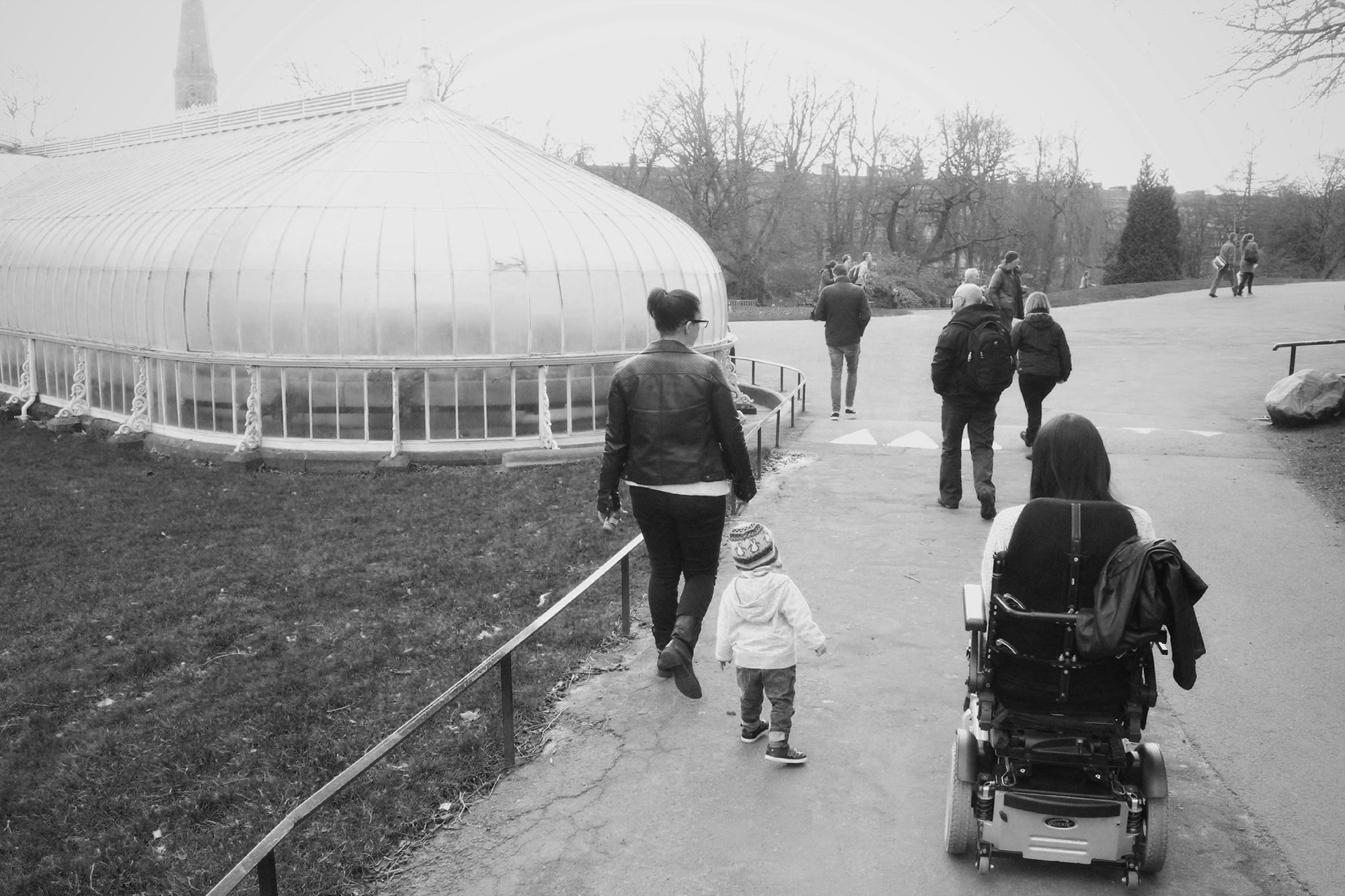 Emma in her power wheelchair, her sister and nephew walking beside her. They have their backs to the camera heading towards the Glasgow Botanic Gardens in the background.