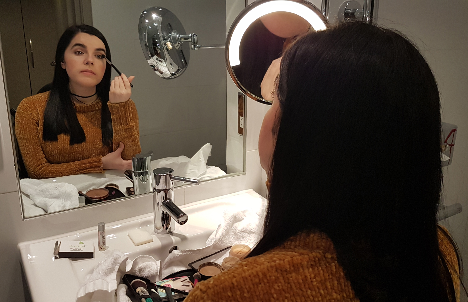 Emma of Simply Emma doing her makeup in her hotel room wearing a mustard chenille jumper.