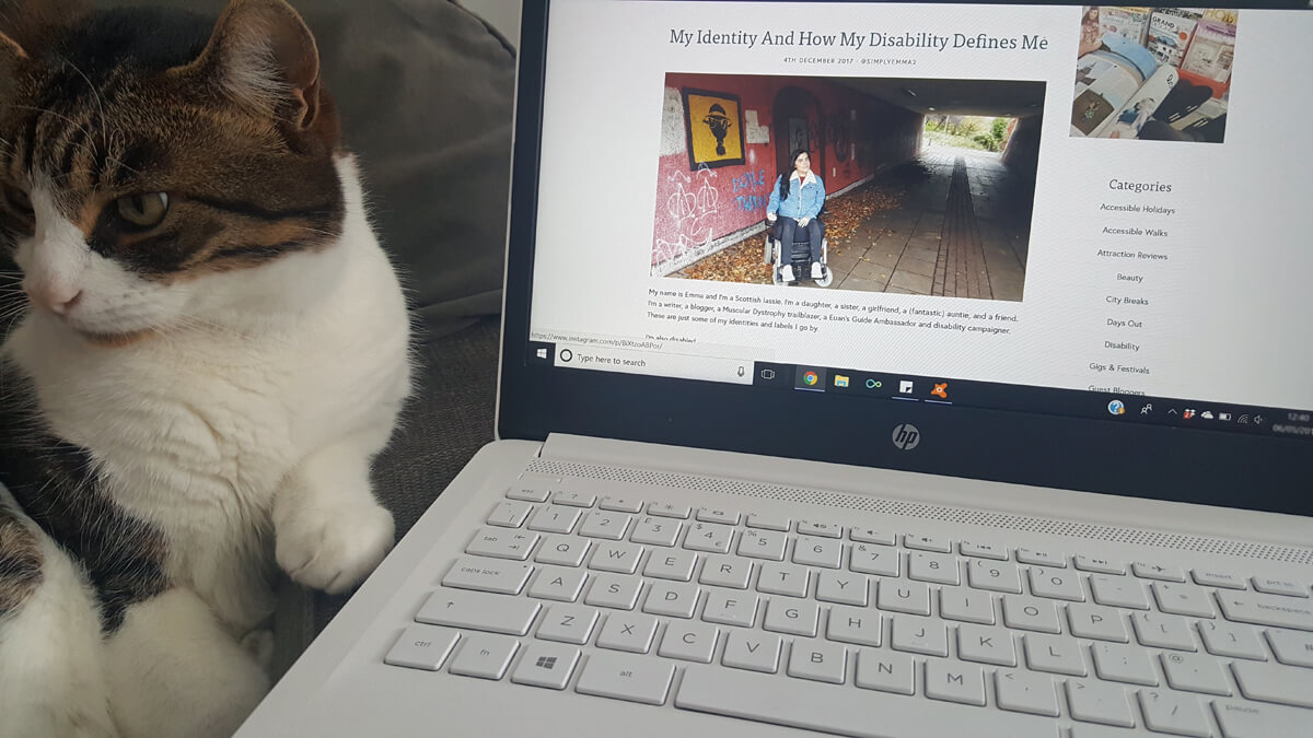 Photo of my laptop with my blog on the screen. It's focusing on my blog post titled 'My Identity and how my disability defines me'. My cat Milo is sitting beside the laptop.