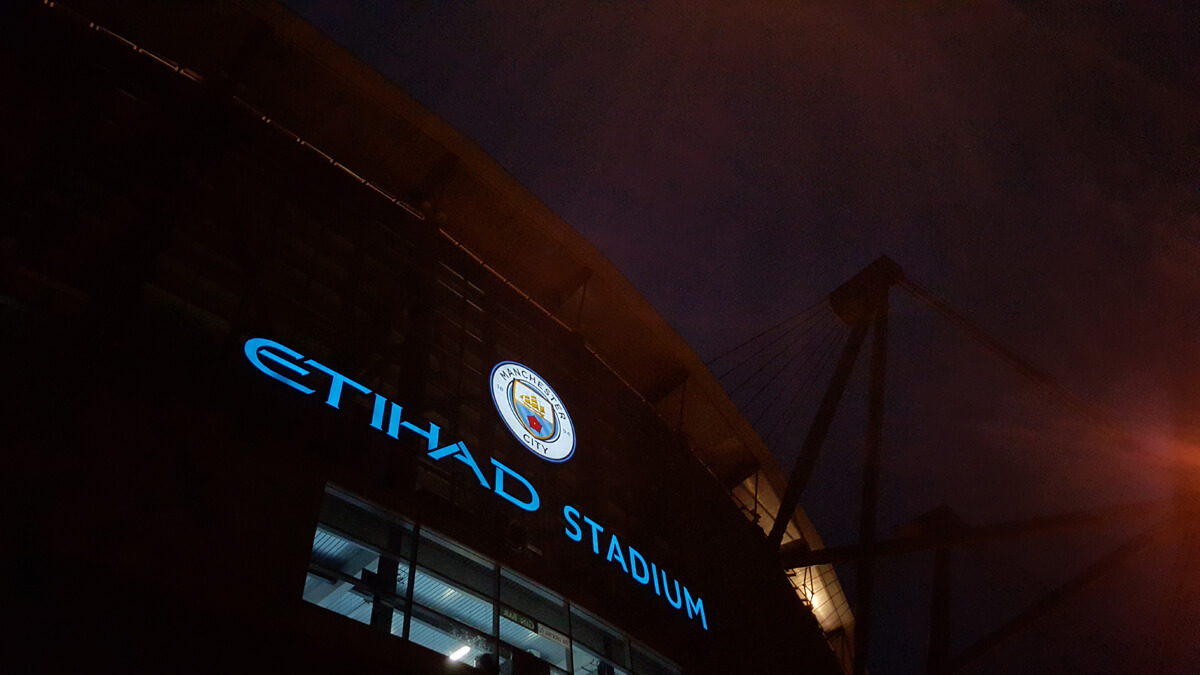 The Etihad Stadium sign on the front of the building taken at night. The sign is lit up.
