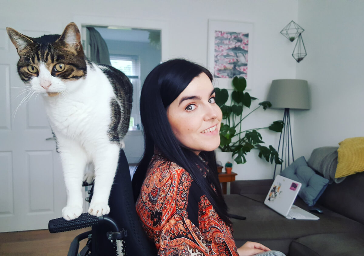 Emma is wearing a paisley pattern shirt and her cat, Milo is standing on the back of her wheelchair.