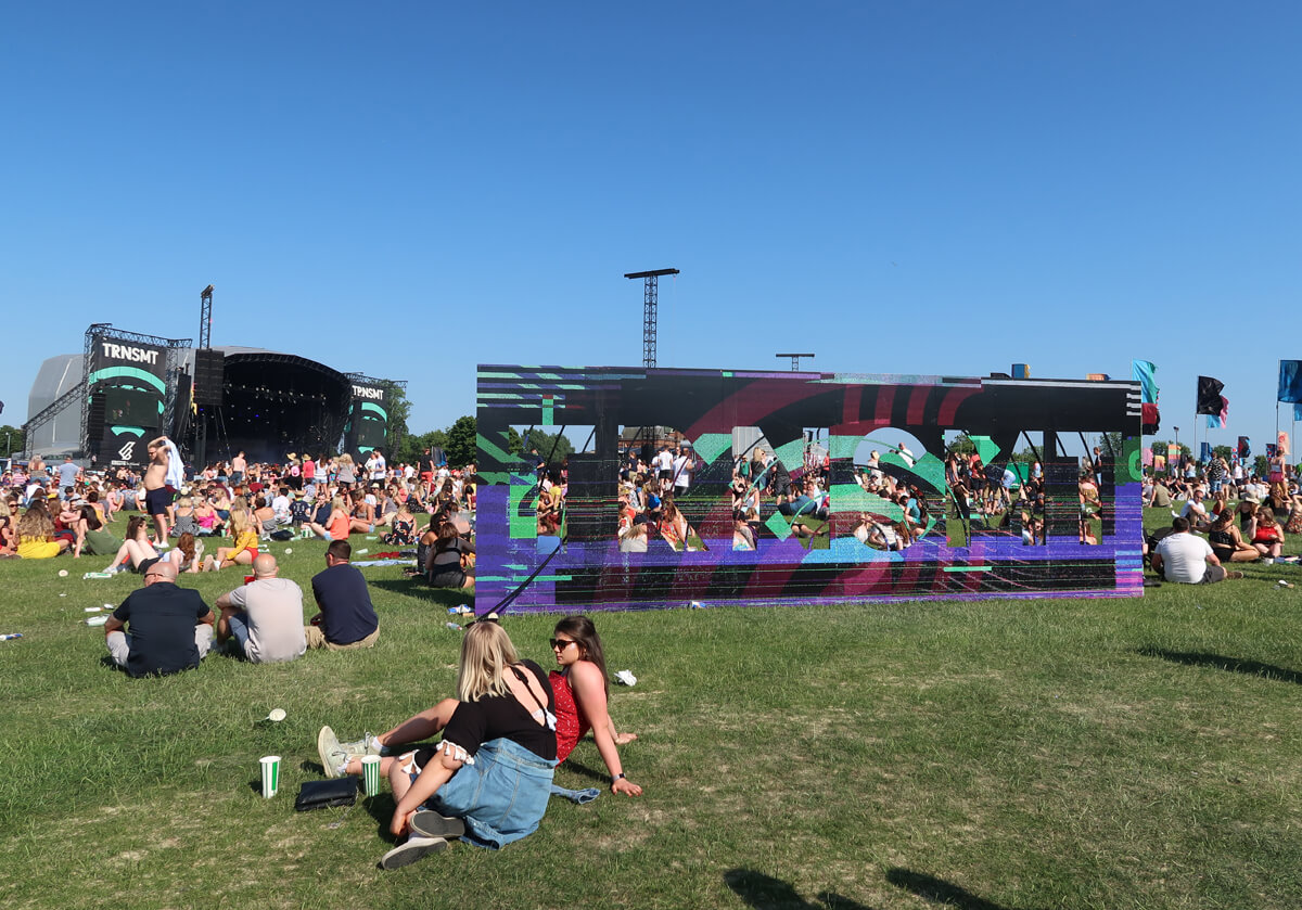 The TRNSMT festival sign in the middle of the festival grounds. There are people sitting on the grass. The main stage is in the background.