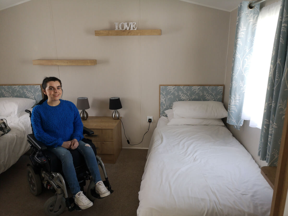 Emma sitting in her powered wheelchair in the wheelchair accessible bedroom. She is sitting inbetween the twin beds. Emma is wearing a blue jumper, jeans and white converse shoes.