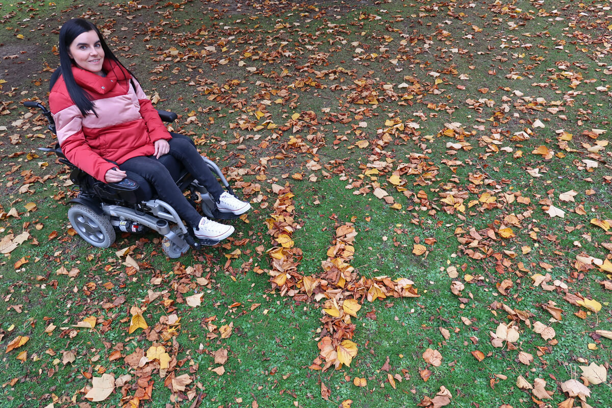 Emma is wearing a pink/red puffy jacket, black jeans and white converse shoes while sitting in her powered wheelchair. She is in her local park. She is sitting beside a giant number four shaped out of leaves.
