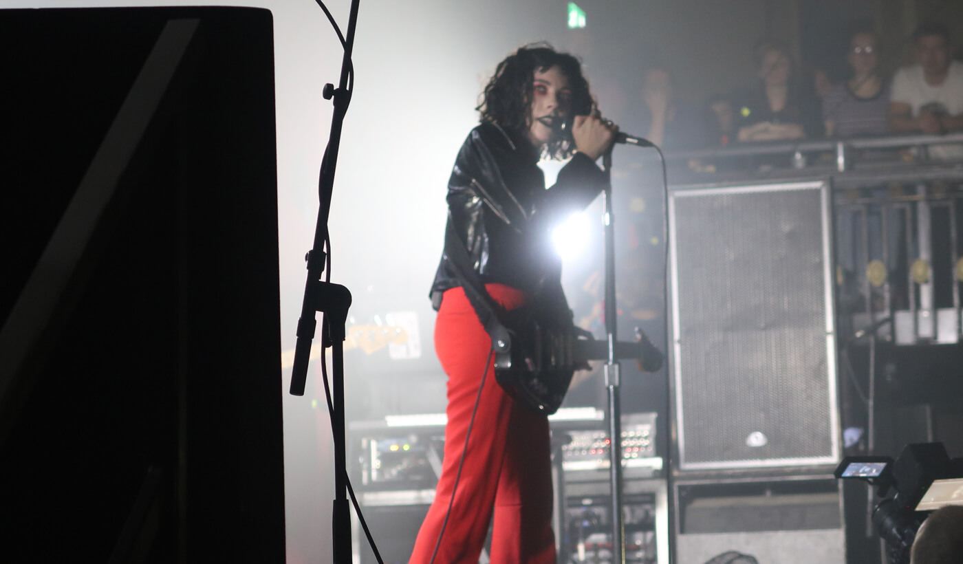 The singer of Pale Waves, Heather is standing on stage singing into the mic wearing bright red trousers and a black leather jacket. She is playing guitar and has red eyeshadow.