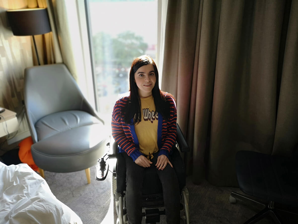 Emma is sitting in her hotel room wearing a yellow t-shirt with the words 'free wheelin' and a blue and orange stripey cardigan. She is looking at the camera smiling.