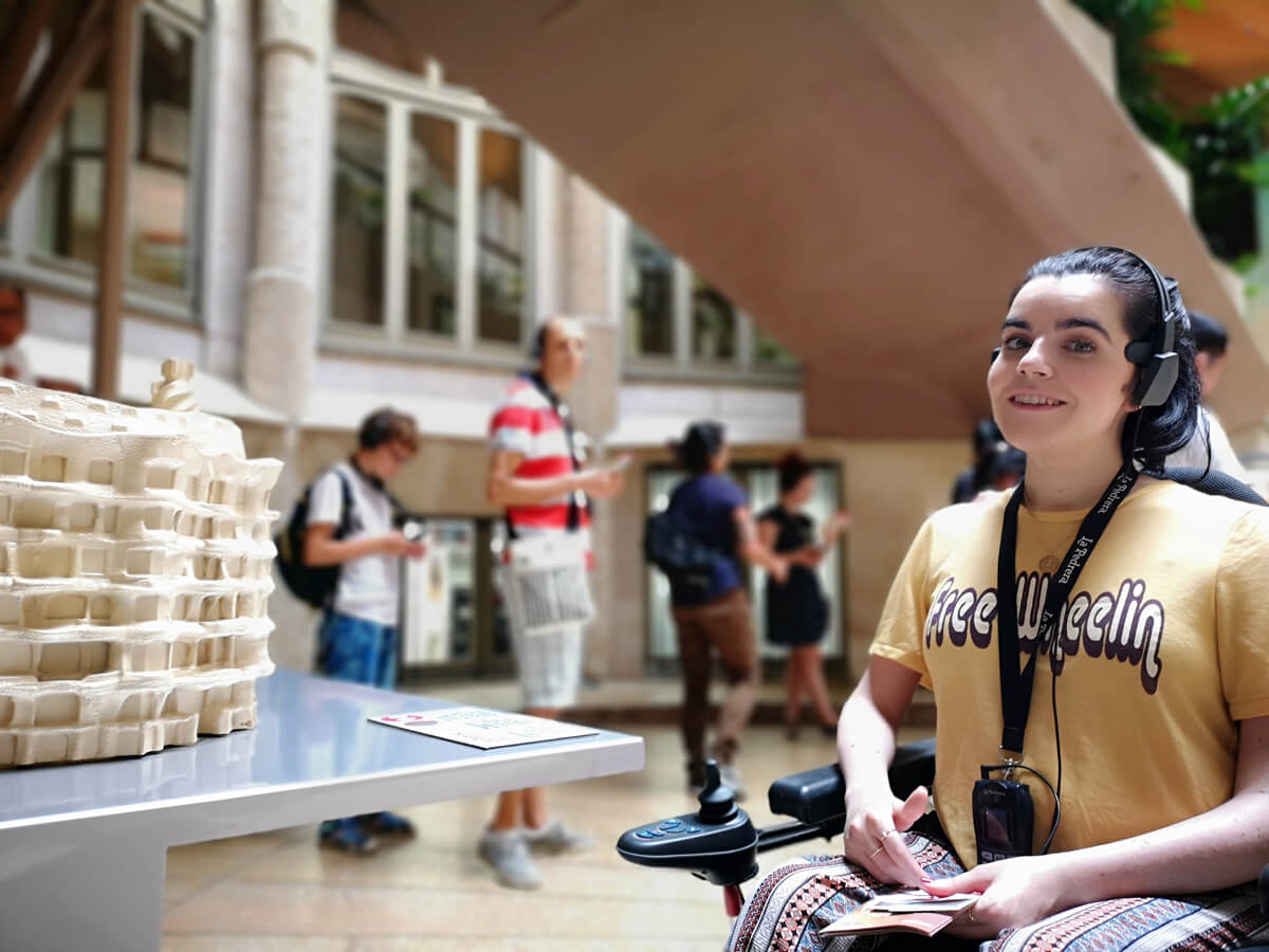 Emma is sitting in her wheelchair wearing a yellow t-shirt with the words 'Free Wheelin'. She is wearing an audio set for the tour of Casa Milà.