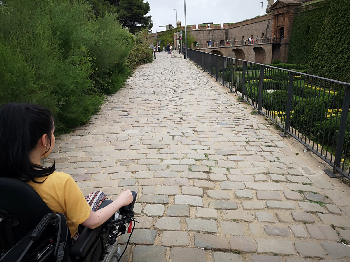 Emma driving her wheelchair along a cobblestone path towards Montjuïc Castle.