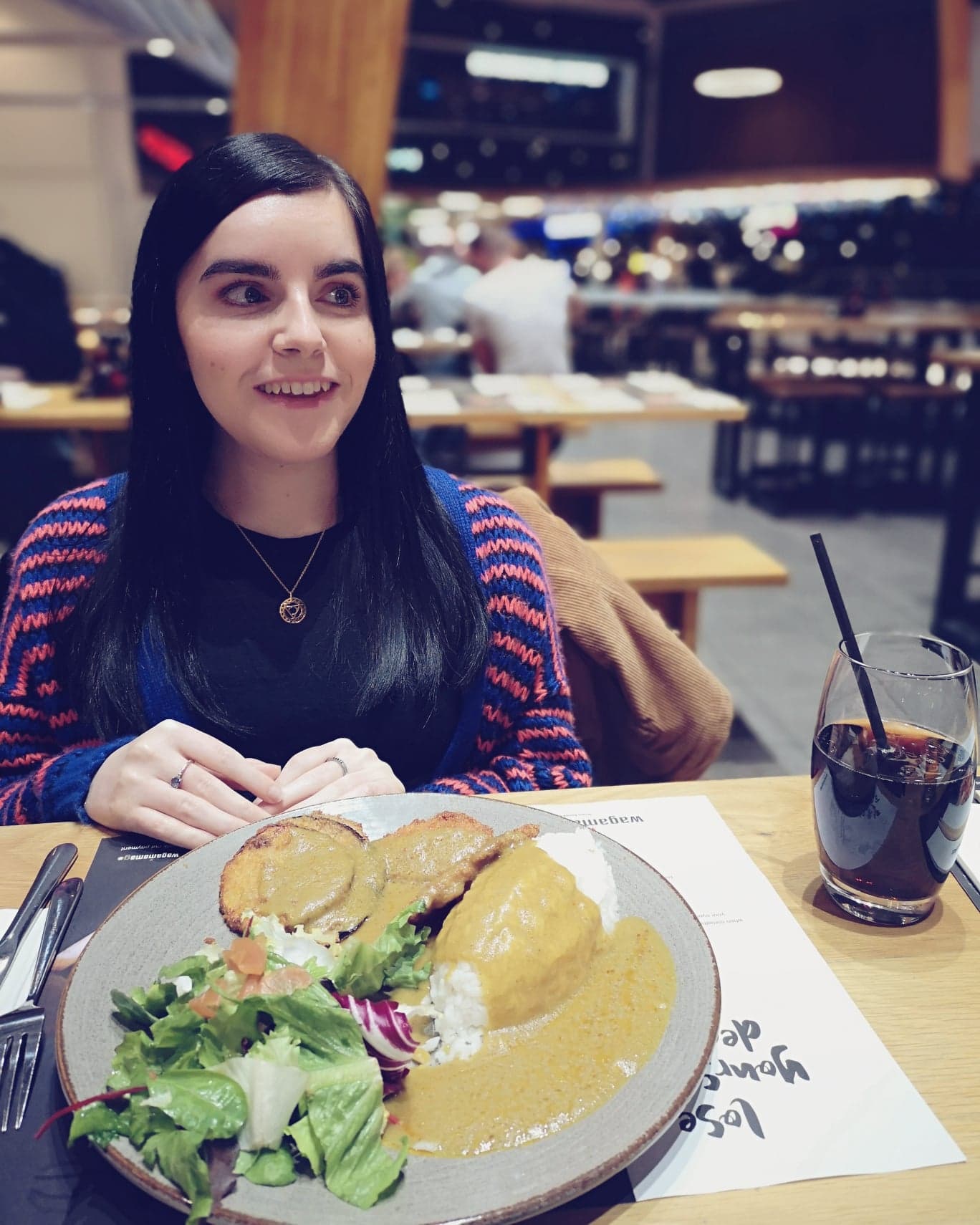 Emma is sitting in Wagamama with a vegan curry. She is wearing a blue and orange stripe cardigan and a black top. She is looking off to the side smiling.