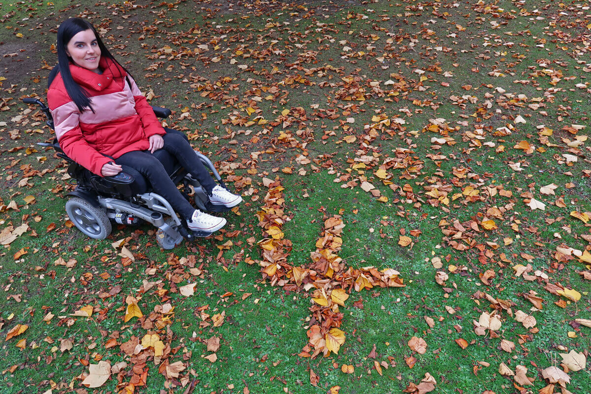 Emma is wearing a pink/red puffy jacket, black jeans and white converse shoes while sitting in her powered wheelchair. She is in her local park. She is sitting beside a giant number four shaped out of leaves.