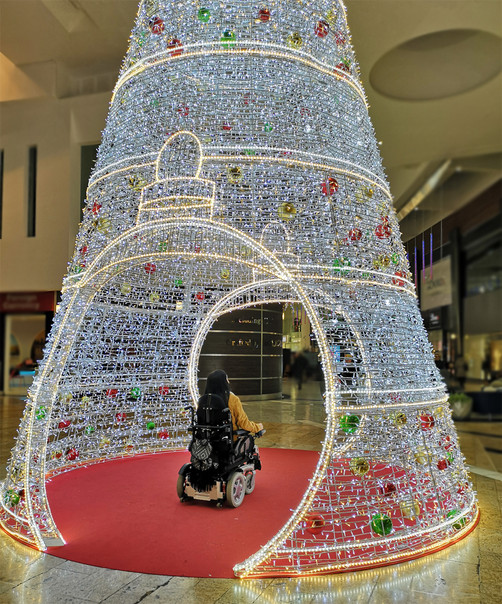 Emma in her powered wheelchair sitting inside a decorative Christmas tree and Christmas light display in Silverburn Shopping Centre.
