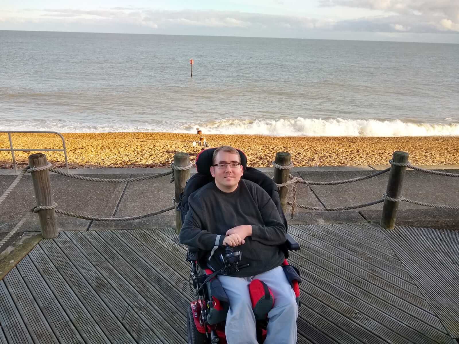 Alex Squire sitting in his wheelchair on the cottage decking. The beach is in the background.