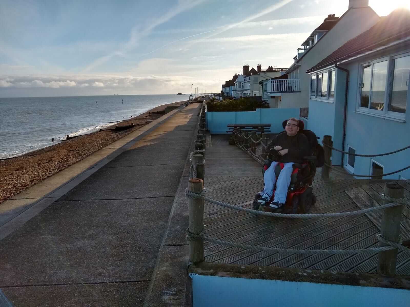 Alex sitting in his powered wheelchair on the cottage decking admiring the view of the beach.