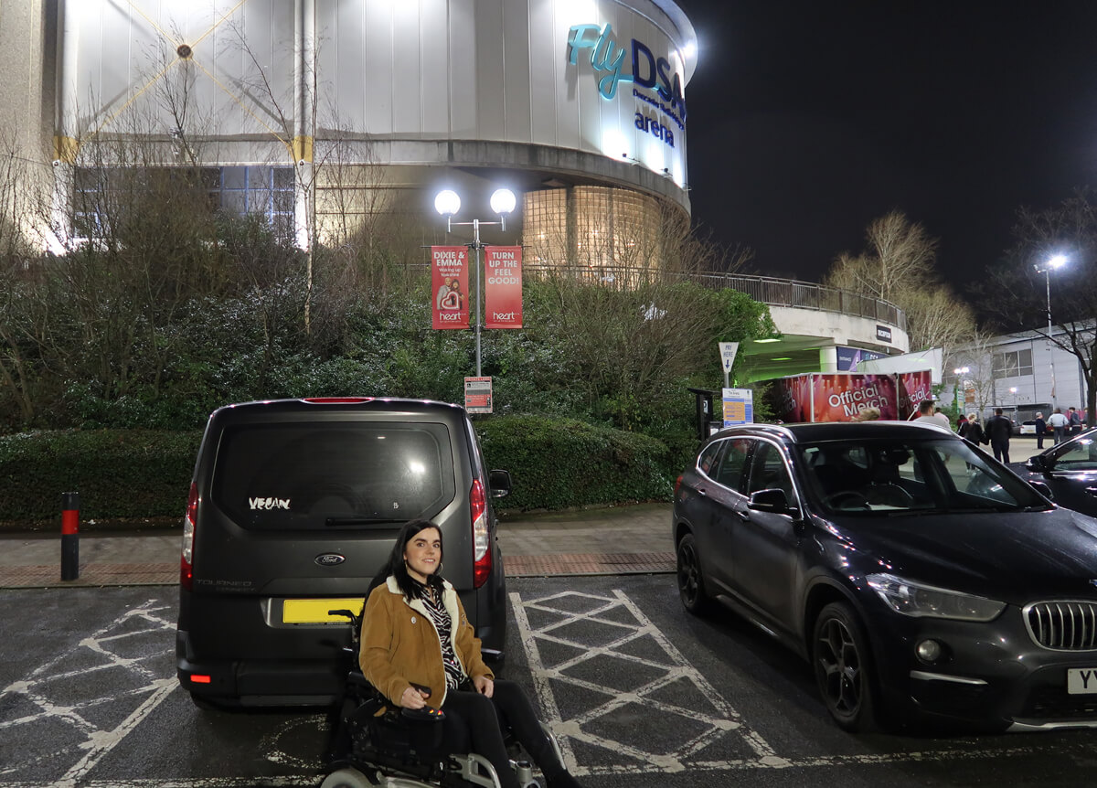 Emma in her wheelchair sitting beside her car in a disabled parking bay at FlyDSA Arena Sheffield.