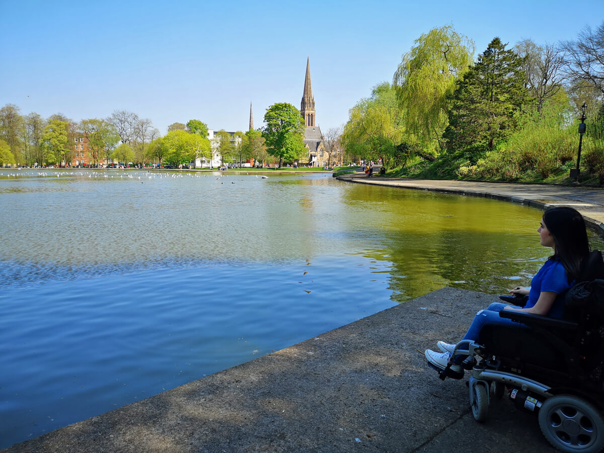 Emma sitting in her wheelchair looking across the pond in Queen's Park.