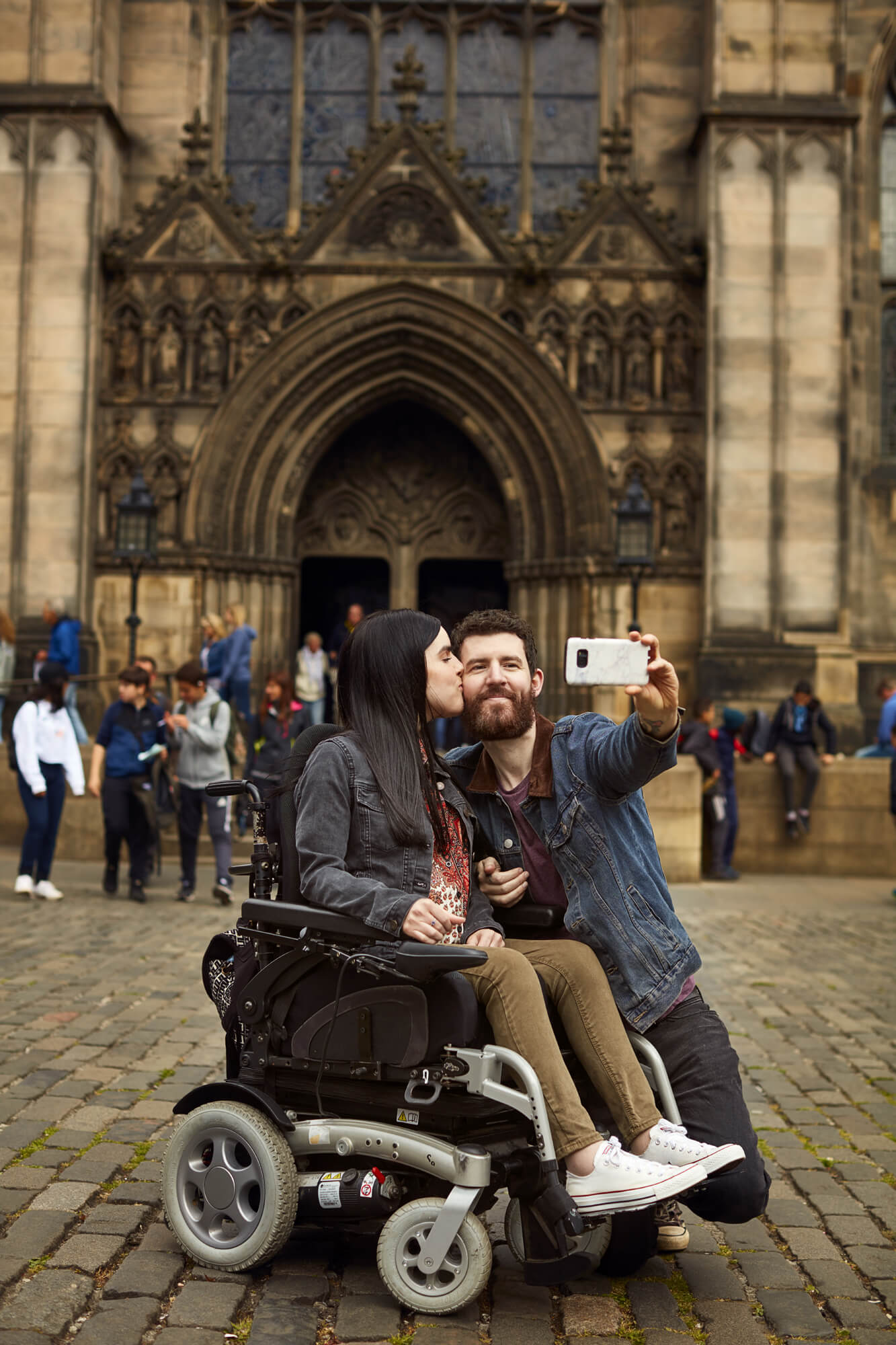 Emma in her powered wheelchair with her boyfriend kneeling beside her holding a phone up while taking a selfie of them together. Emma is giving him a kiss on the cheek. They are outside a Cathedral in Edinburgh.