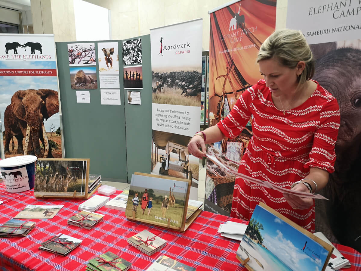 A woman standing behind the merch and ticket table. She is looking through the tickets for ours. She is wearing a red dress. There are photographs of Elephants hanging up around her.