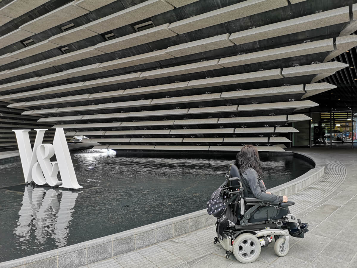 Emma in her wheelchair with her back to the camera. She is facing the entrance of the V&A while sitting beside the swallow water surrounding the building. There are large white letters "V&A" sitting in the water.