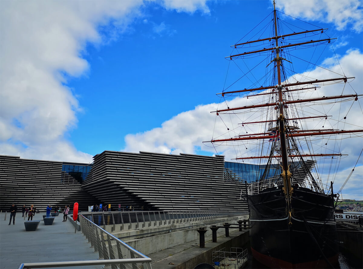 Exterior shot of the V&A Dundee against a blue sky with white fluffy clouds. The RRS Discovery ship is sitting beside V&A on the Dundee Waterfront.