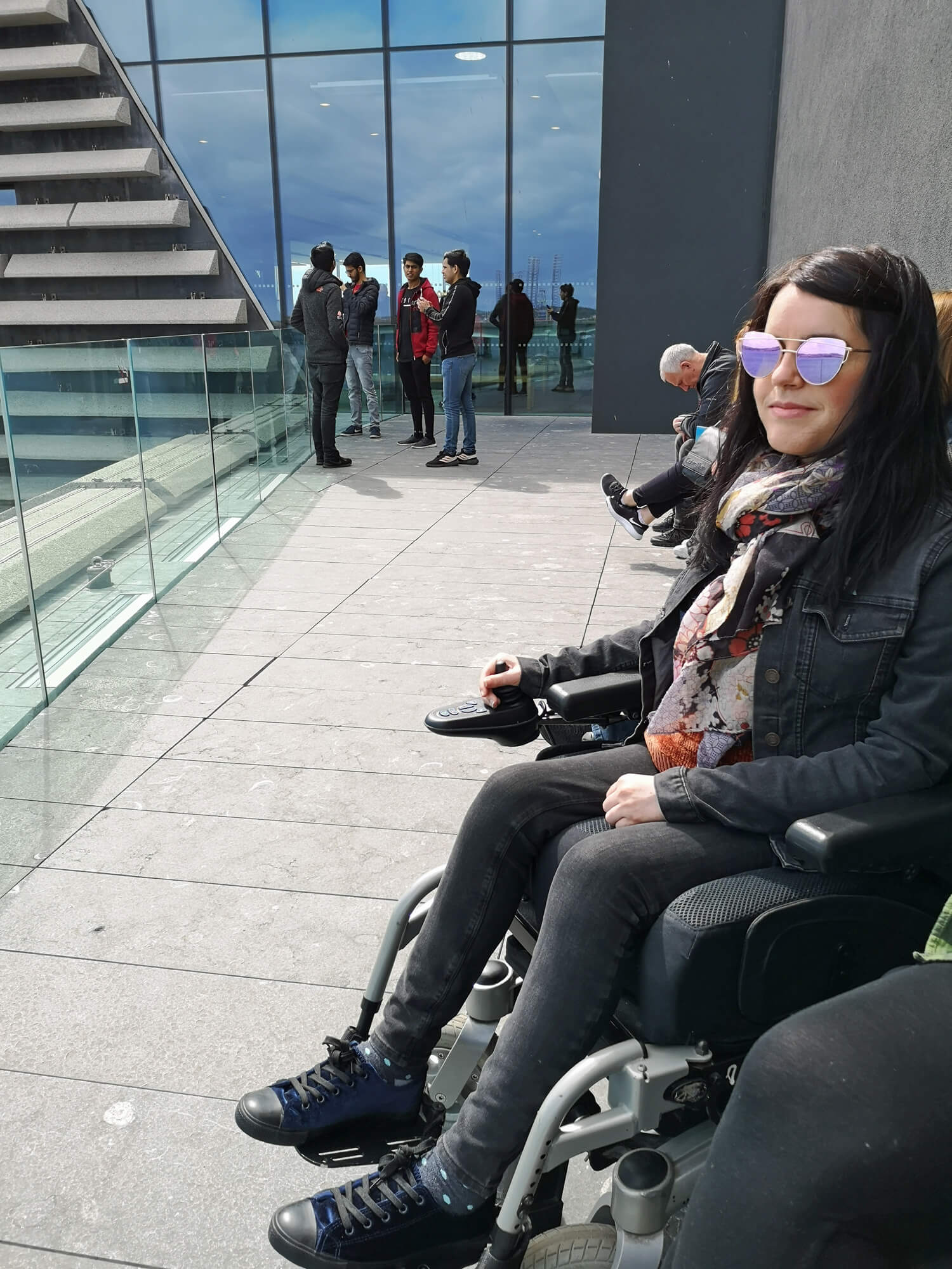 Emma sitting on the viewing terrace. She is wearing sunglasses, black denim jacket and black skinny jeans.