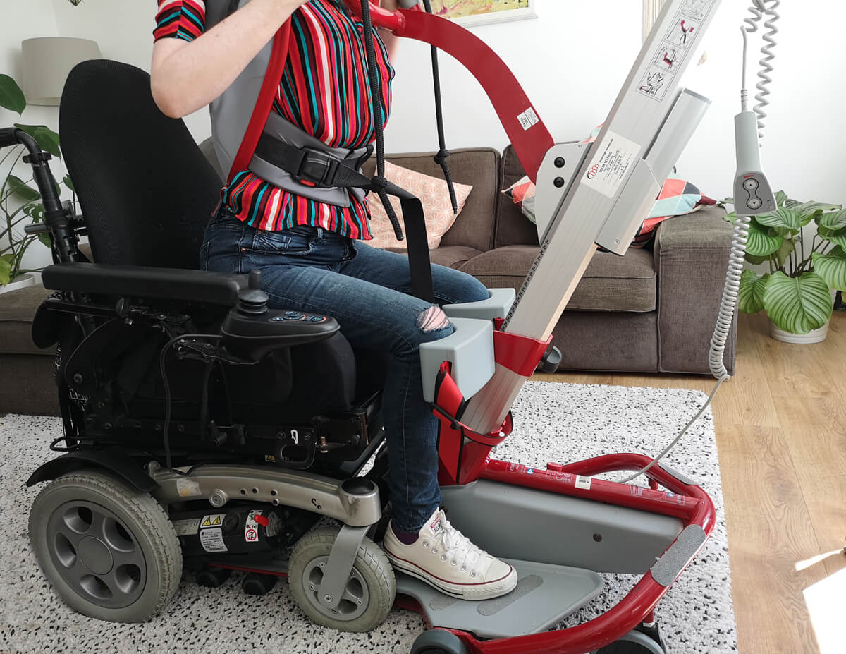 Emma being hoisted from her wheelchair using a Molift Quick Raiser sit-to-stand hoist. She is wearing a multicoloured striped shirt and is in her livingroom.