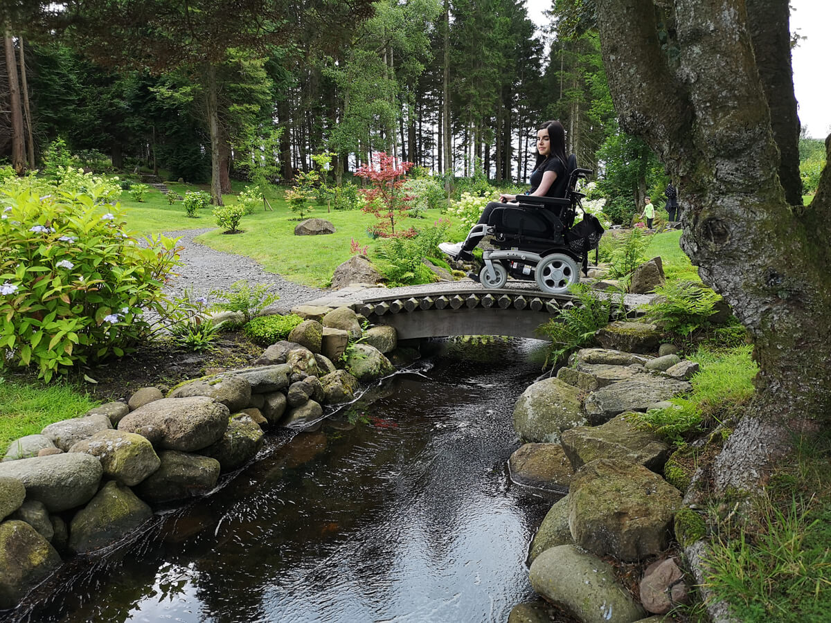 Emma crossing a small wooden bridge in her wheelchair with a stream flowing underneath.