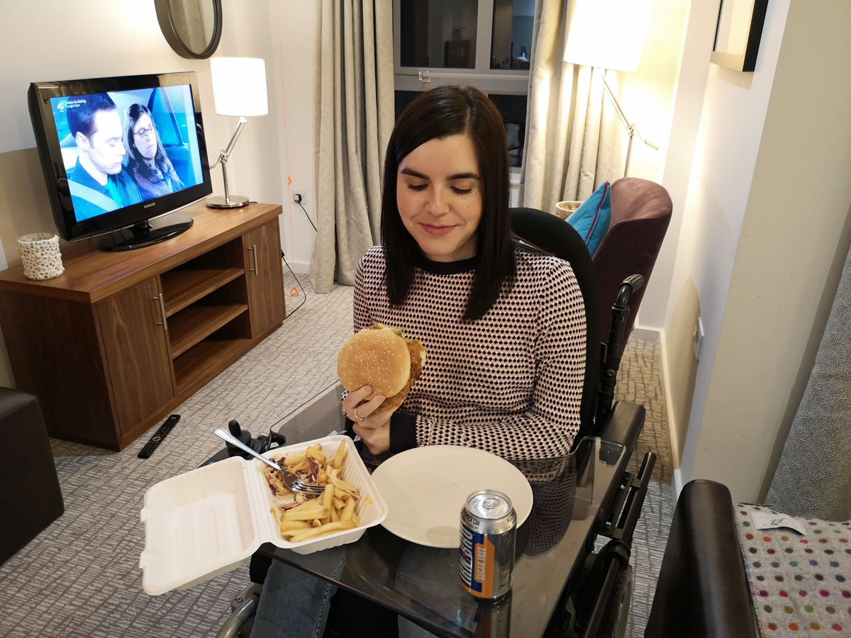 Emma sitting in her wheelchair holding a burger. There is a box of fries and a can of diet Irn Bru next to her on her wheelchair tray table.
