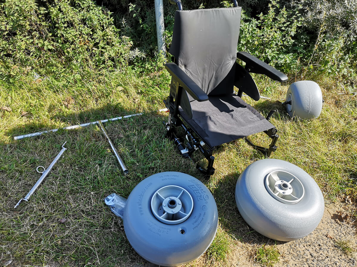 Manual wheelchair sat on the grass with front and back wheels removed. The conversion kits wheels and bars are laid next to the chair on the grass.