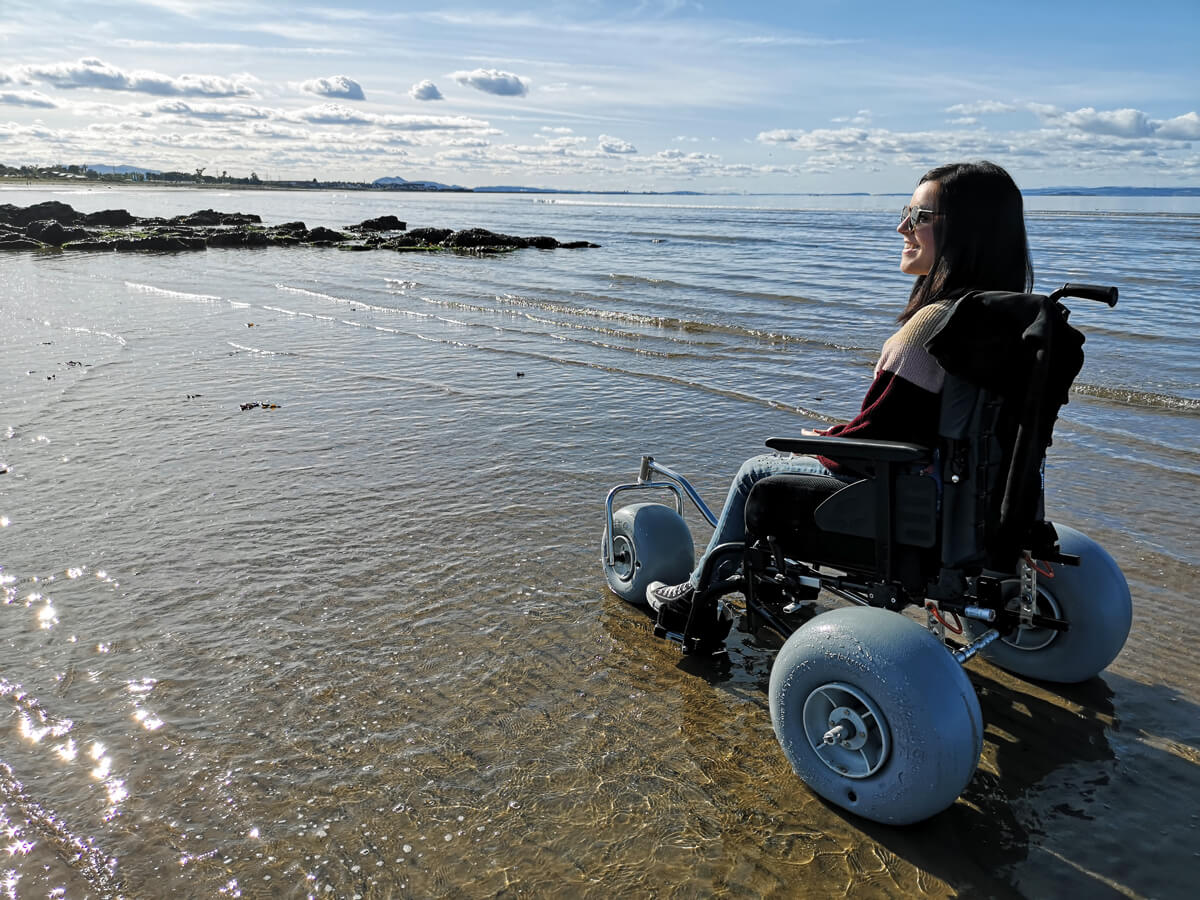 Emma using the WheelEEZ® Beach Wheelchair Conversion Kit at the beach in Scotland. Emma is sitting side on to the camera with her face slightly facing the camera. She is smiling and wearing sunglasses. She is sitting in her manual wheelchair with the conversion kit attached. She is sitting in the water a little bit.