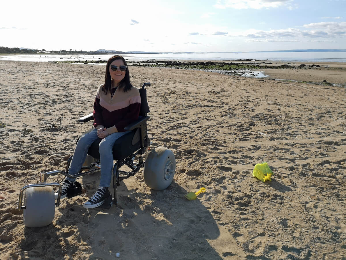 Emma using WheelEEZ® Beach Wheelchair Conversion Kit on the beach. She is smiling. The water is behind her and a bucket for building sandcastles is on the sand beside her.