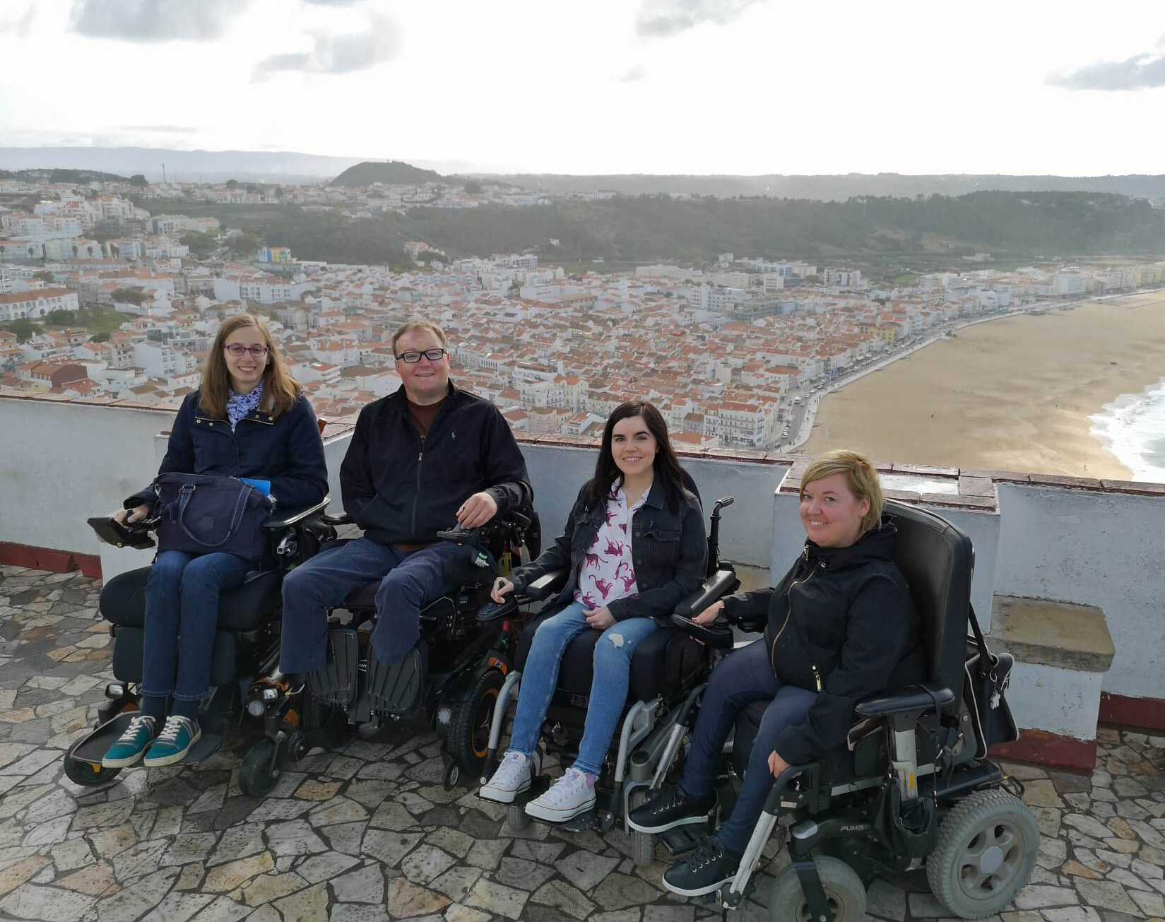 Emma with John Morris, Sanna and Blandine, a group of accessible travel bloggers. They are sitting in their wheelchairs with the Nazaré beach behind them.