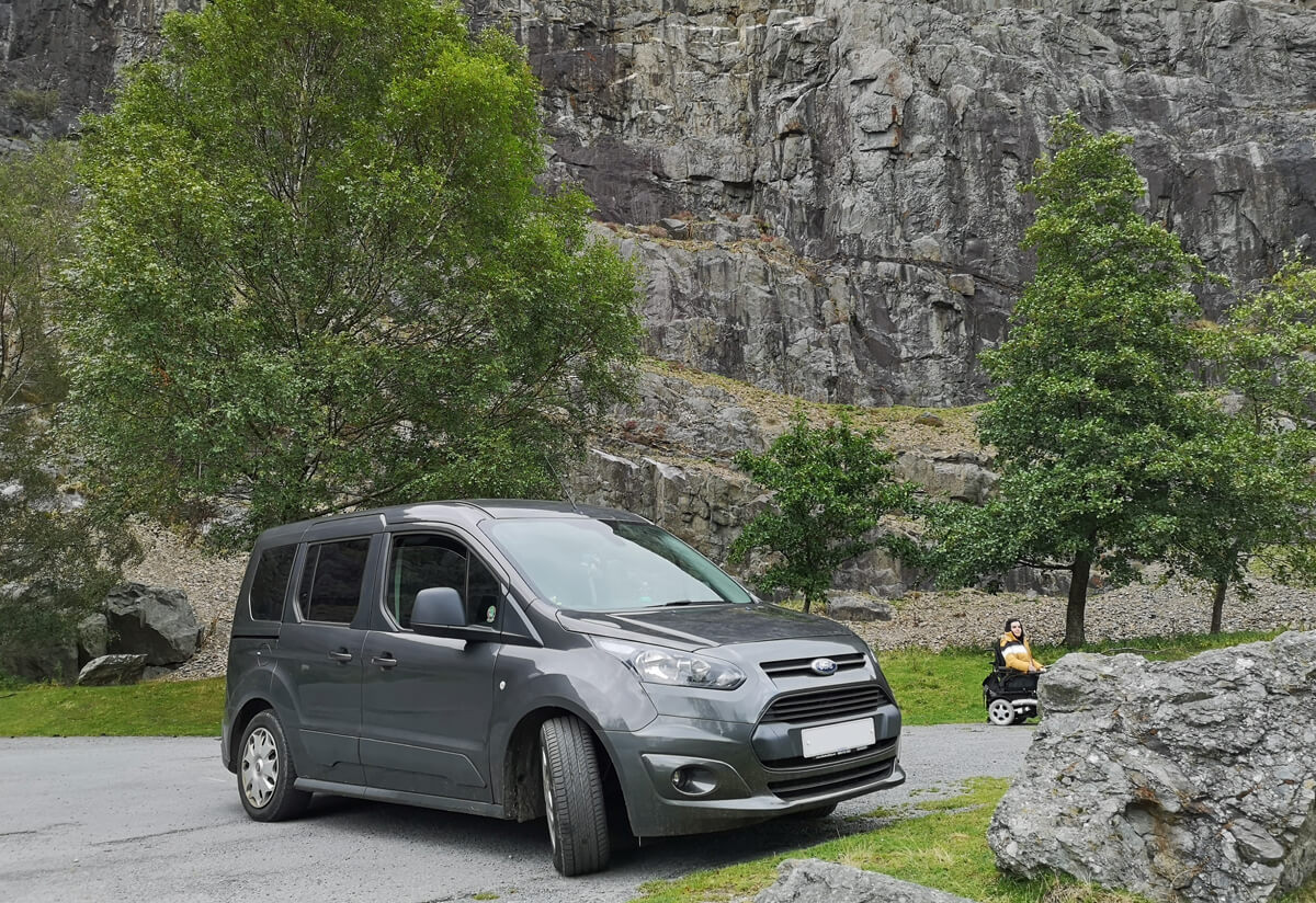 A Ford Tourneo Connect wheelchair accessible vehicle parked beside a rock wall. Emma is sitting beside the vehicle too.