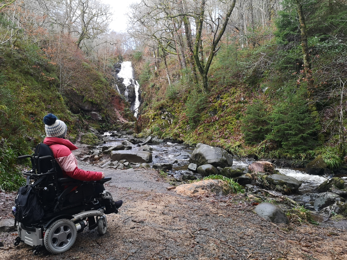 Emma in her wheelchair looking across a river to the waterfall.