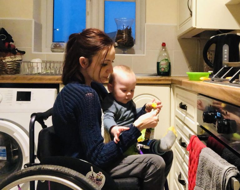 Molly is sitting in her manual wheelchair in her kitchen. Her young son is sitting on her lap.