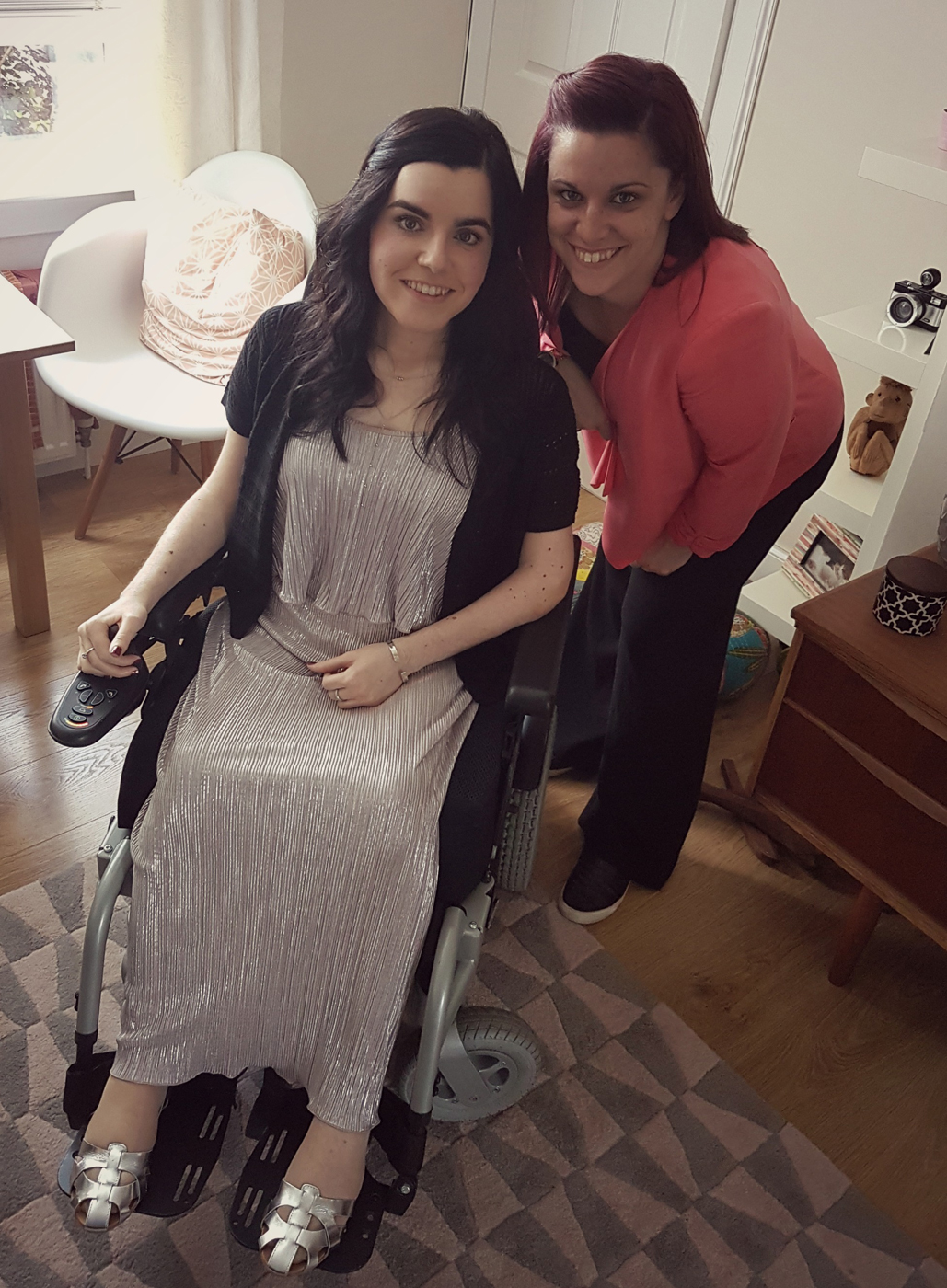 Emma is sitting in her wheelchair. She is wearing a dress. Her sister is standing beside her. They are both smiling.