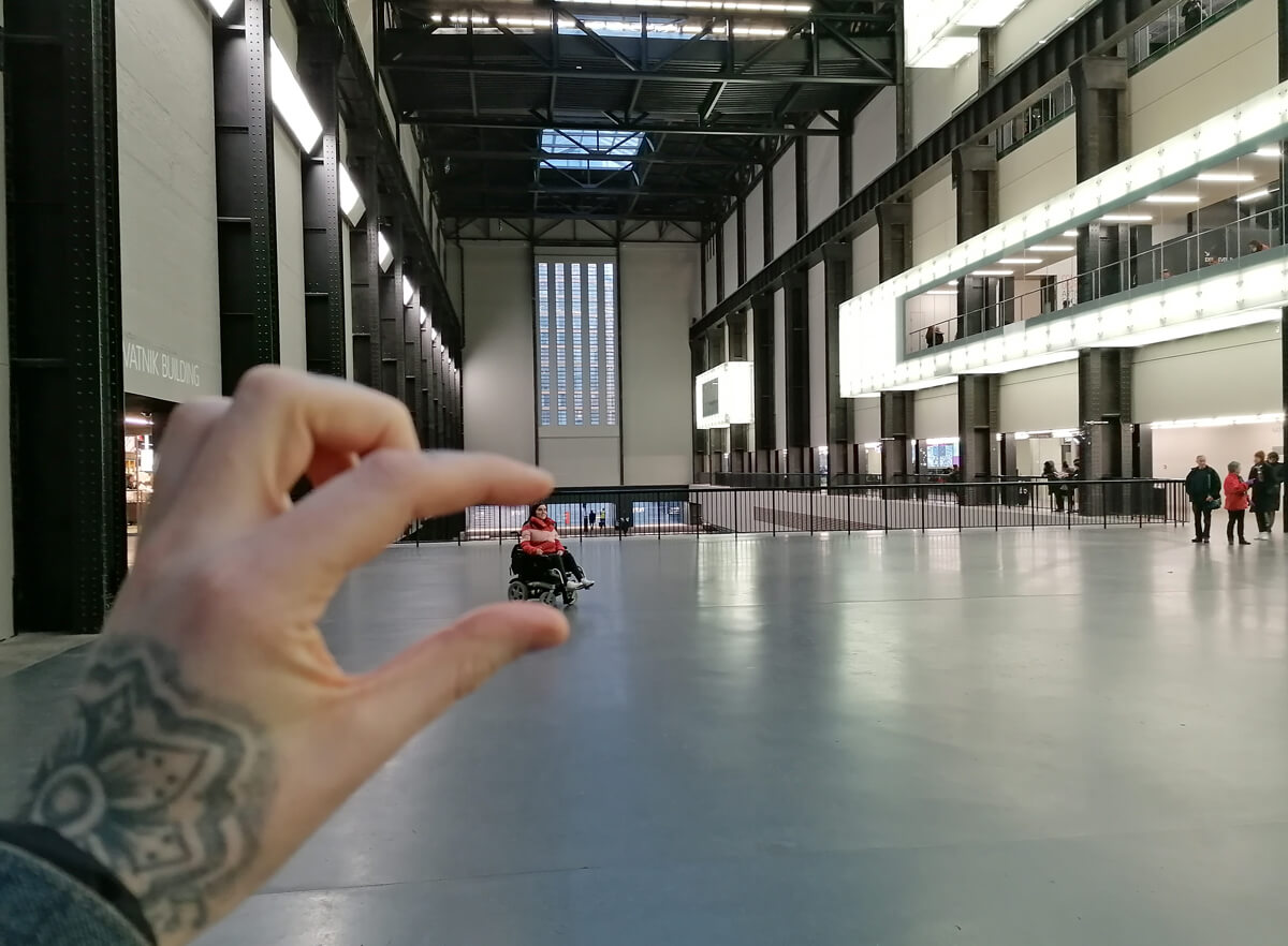 In the forefront of the image is Allan's tattooed hand. He is pinching his fingers together to look like he is squeezing Emma who is way in the distance in the middle of the massive Tate museum.