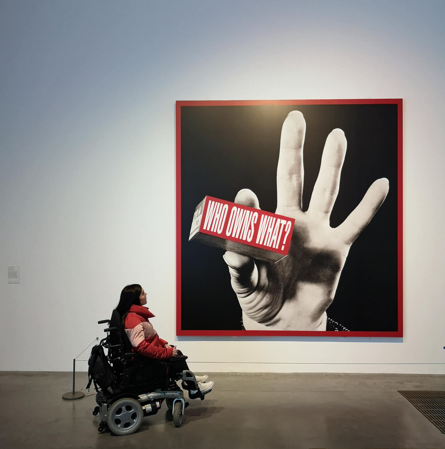 Emma looking at a art piece on the wall with a giant hand holding a sign that says "Who owns what?"