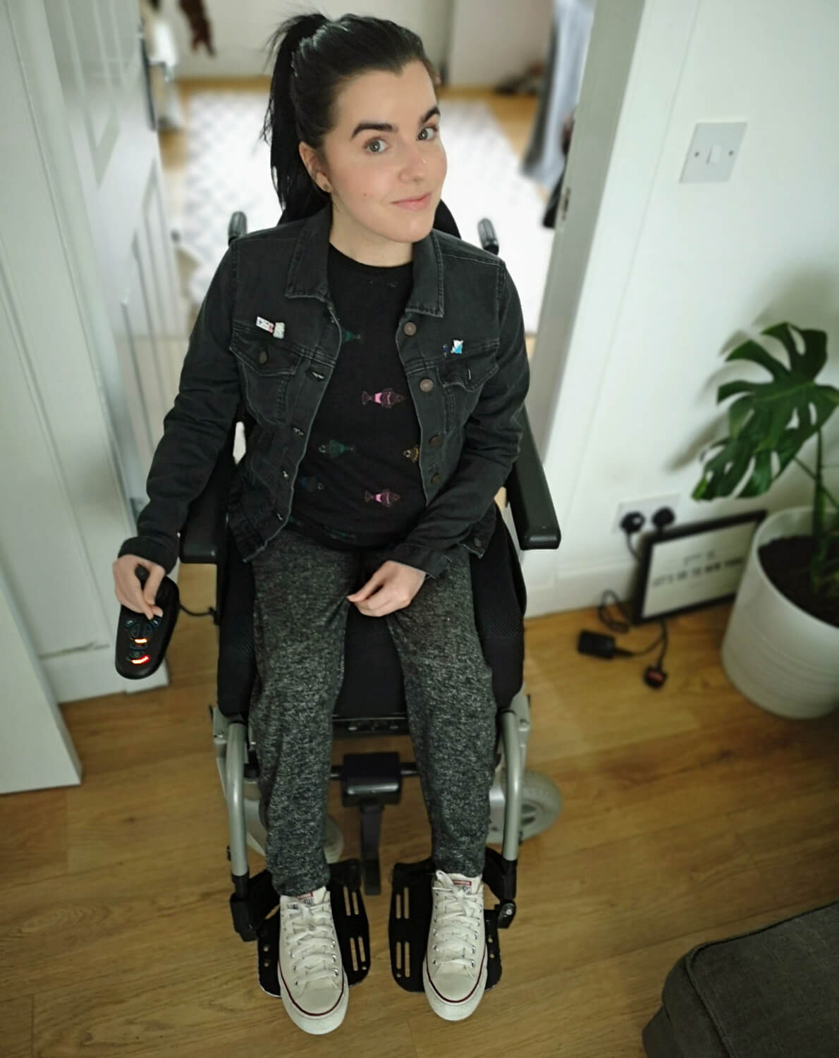 Emma is sitting in her wheelchair in her livingroom. She is wearing grey jogging bottoms, white converse shoes, a black top and black denim jacket.