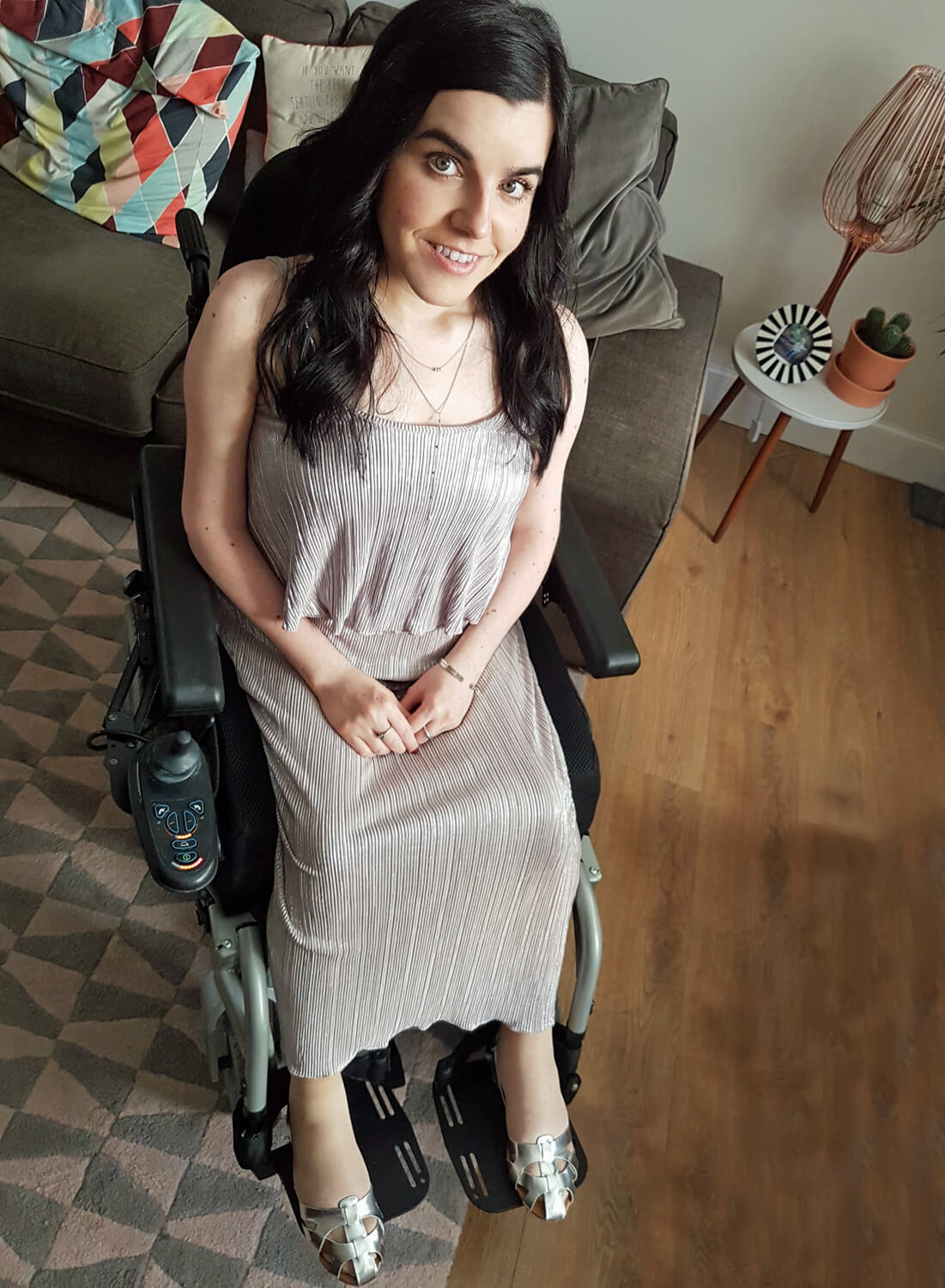 Emma is sitting in her wheelchair wearing a metallic pleated maxi dress. 