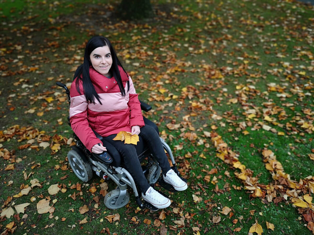 Emma in her wheelchair in the local park. Emma is wearing a red puffy jacket.