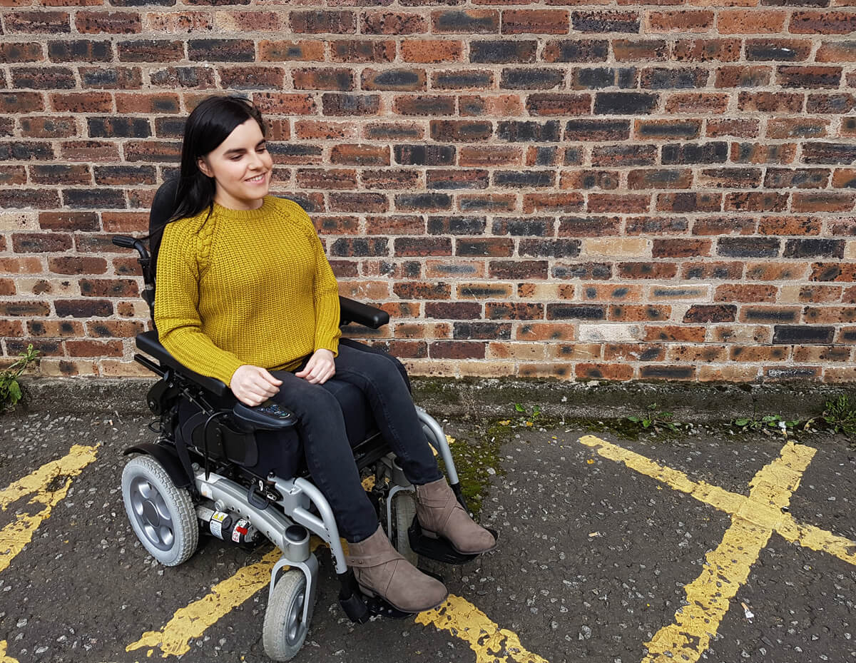 Emma in her wheelchair sitting next to a brick wall. She is wearing brown ankle boots, black skinny jeans and a mustard coloured jumper.
