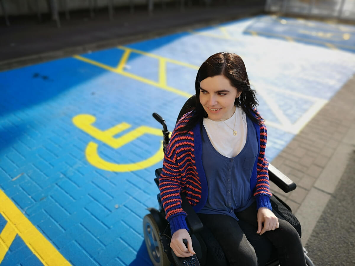 Emma sitting in her wheelchair beside a disabled parking space