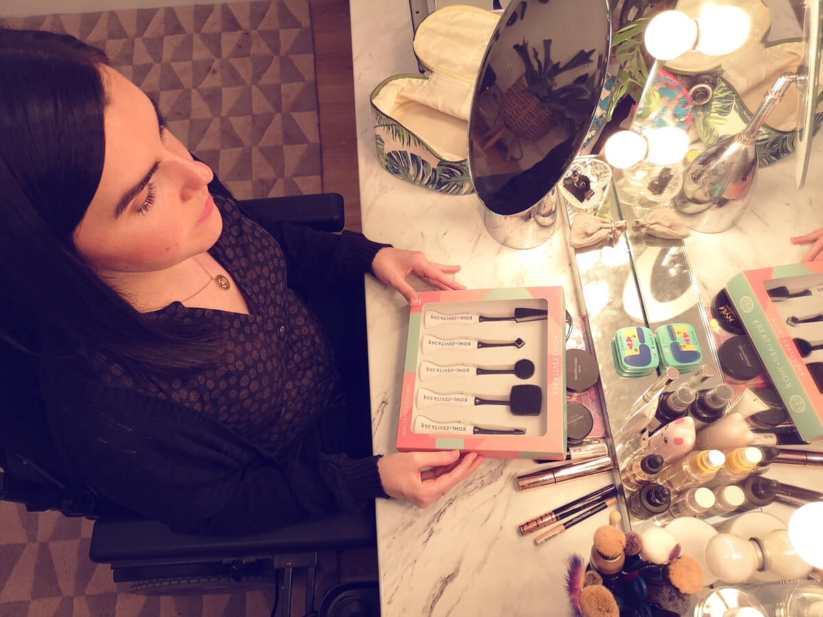 Emma sitting at her makeup vanity table surrounded by her makeup. Emma is holding a box displaying five makeup brushes.