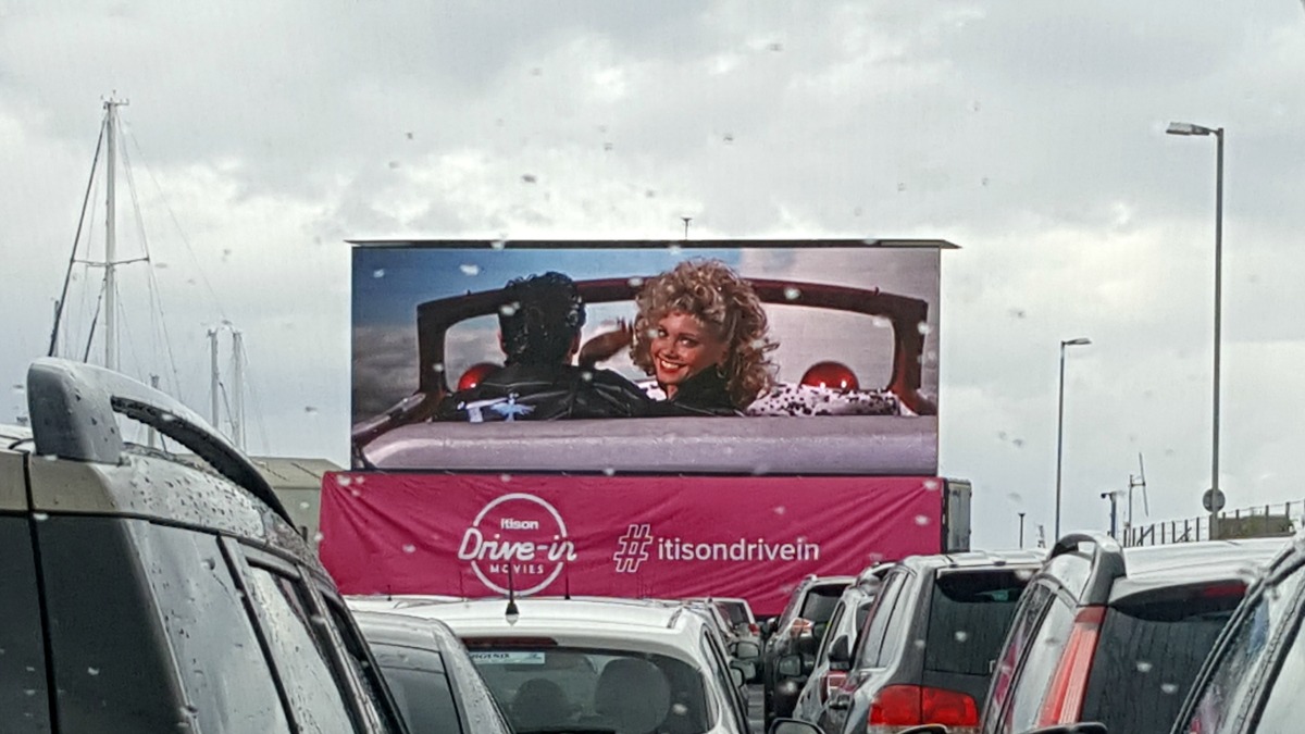 Sandy and Danny from Grease on the large outdoor movie screen. There are rows of parked cars at the bottom of the photo.