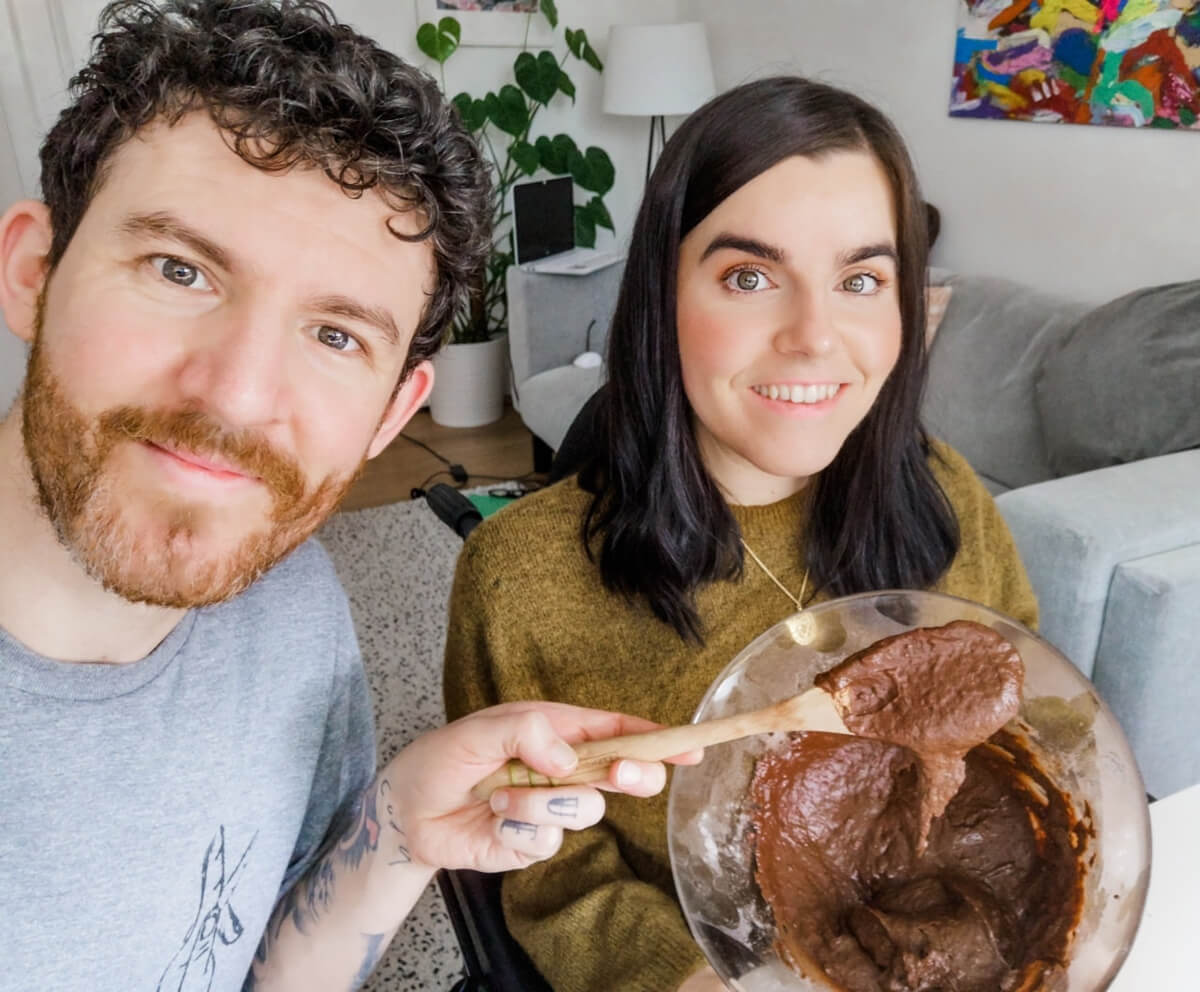 Emma and Allan smiling while holding a bowl of chocolate vegan batter. Allan is holding a wooden spoon.