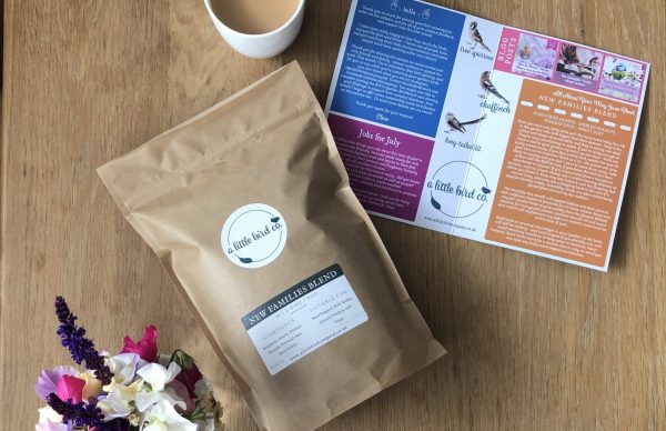 A brown bag of bird food laid on top of a table with a leaflet opened up next to it as well as a coffee and flowers.