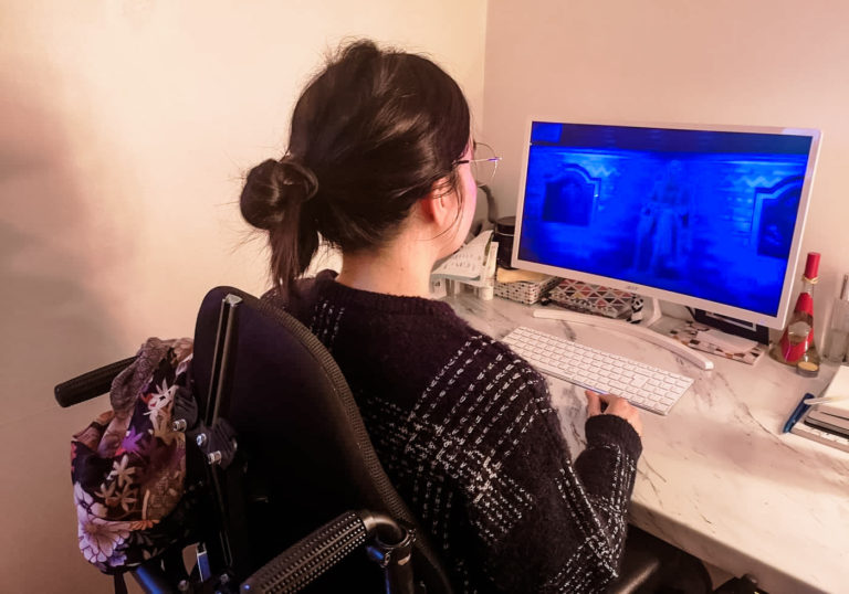 Emma sitting in her wheelchair with her back facing the camera. She is at her desktop PC watching Dermot Kennedy's virtual gig.