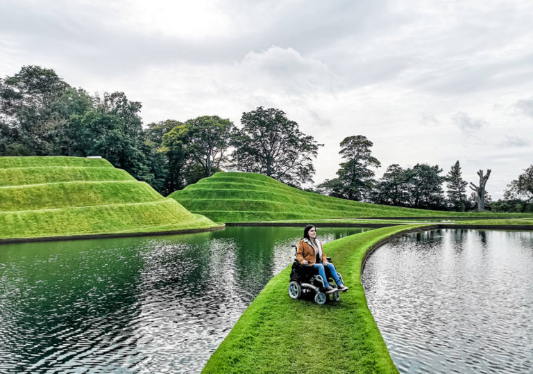Emma is sitting in her powered wheelchair on a strip of grass with water on each side of her. Behind her is giant swirly mounds of grass shaped like pyramids.