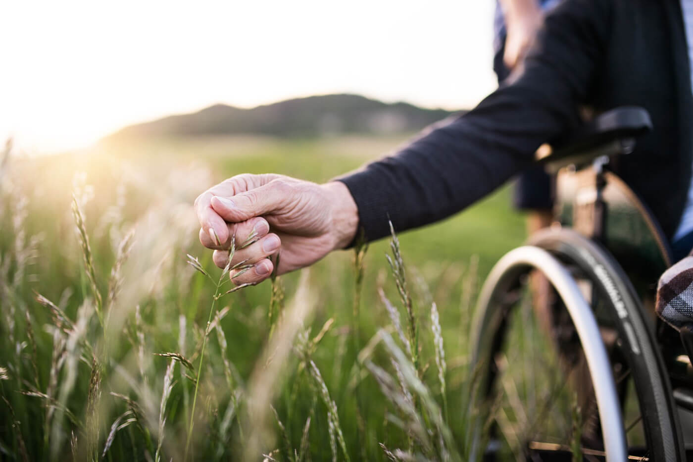 An elderly mans hand reaching out to touch grass while sitting in a wheelchair in the middle of a field.