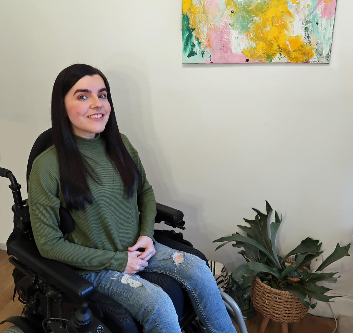 Emma sitting in her powered wheelchair. She has long dark hair. She is wearing a khaki coloured long sleeve top and blue jeans. Emma is smiling at the camera while sitting next to some plants.